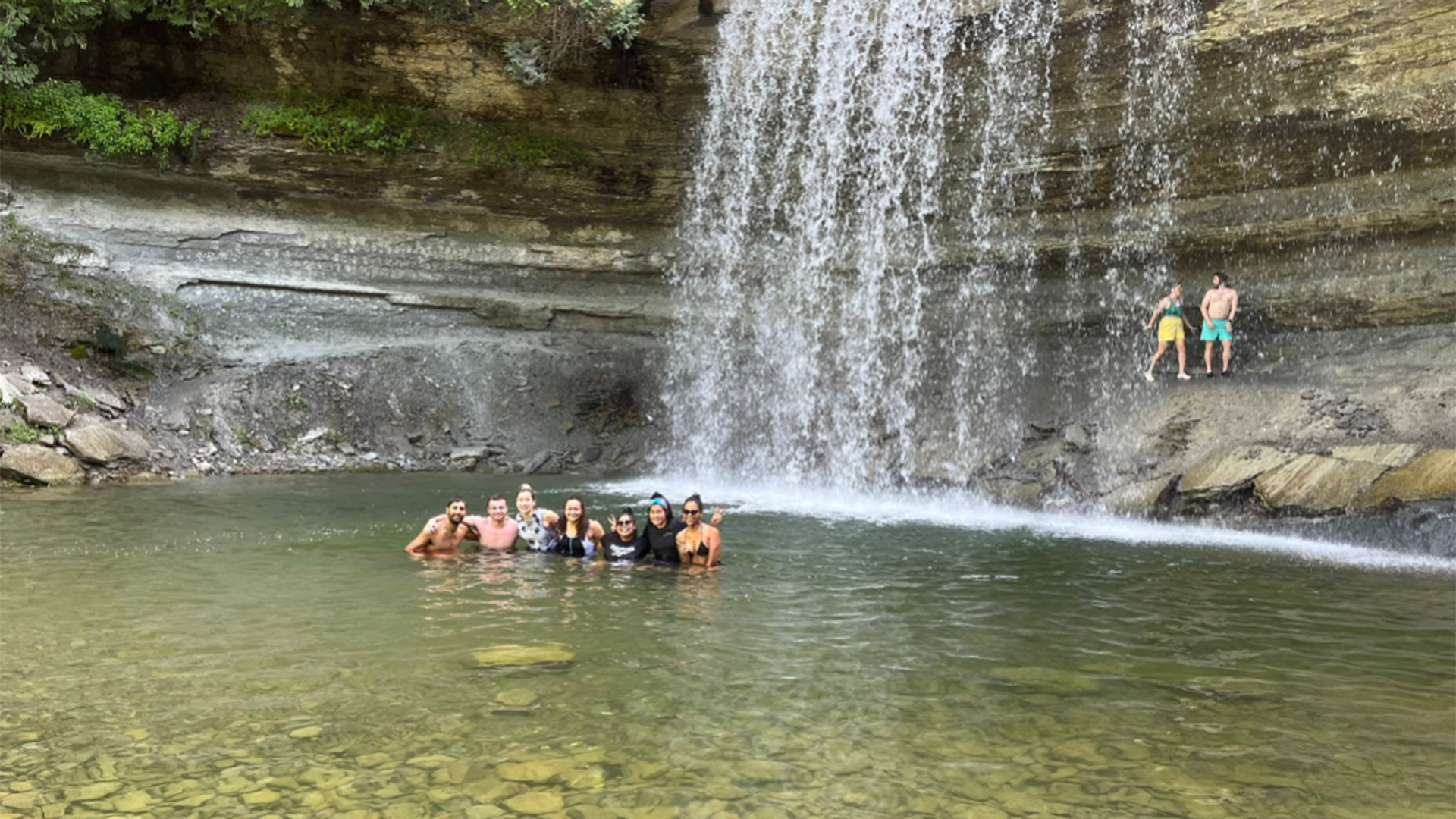 7 people in the middle of a natural body of water huddled together smiling into the camera, under a water fall