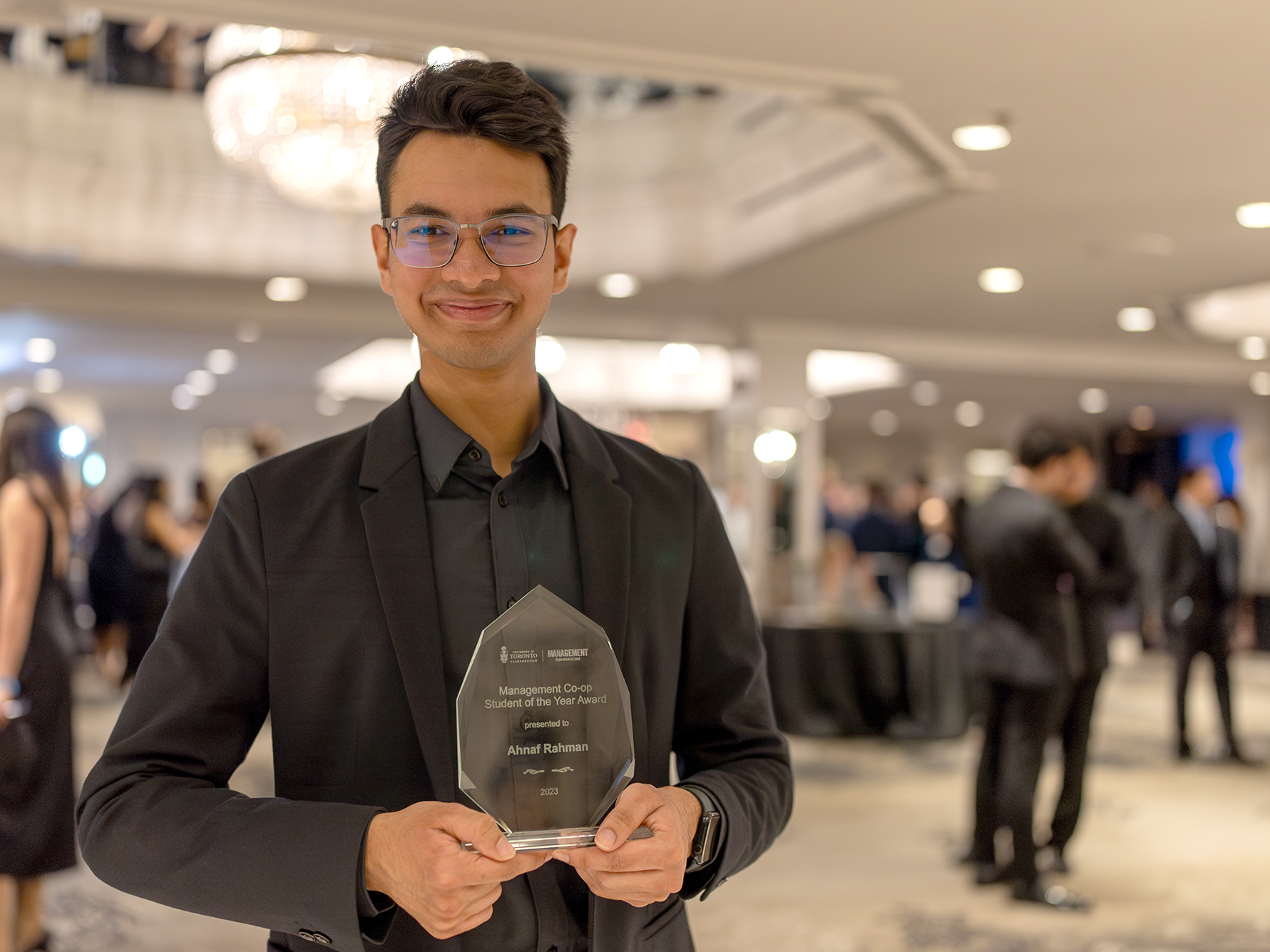 CEWIL Canada Co-op Student of the Year Ahnaf Rahman holds his award and smiles in front of a crystal chandelier while wearing a black suit at the University of Toronto Scarborough Management gala.