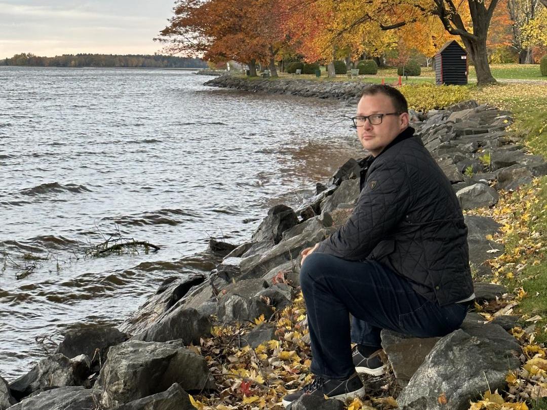Professor Chad Cowie crouching by a rocky lake shoreline in autumn
