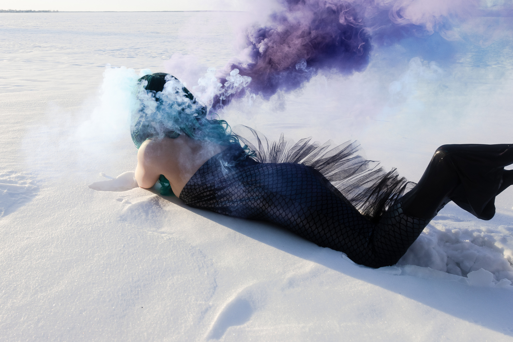 Graphic for the artwork, Sedna | ᓴᓐᓇ #6. On top of the image is a photograph of a woman with long dark green/blue hair lying on her stomach on the snow. The woman’s full body is in frame wearing a mermaid tail with black fabric on the side while her shoulders are exposed. There are plumes of grey, blue, and purple smoke coming from her head.