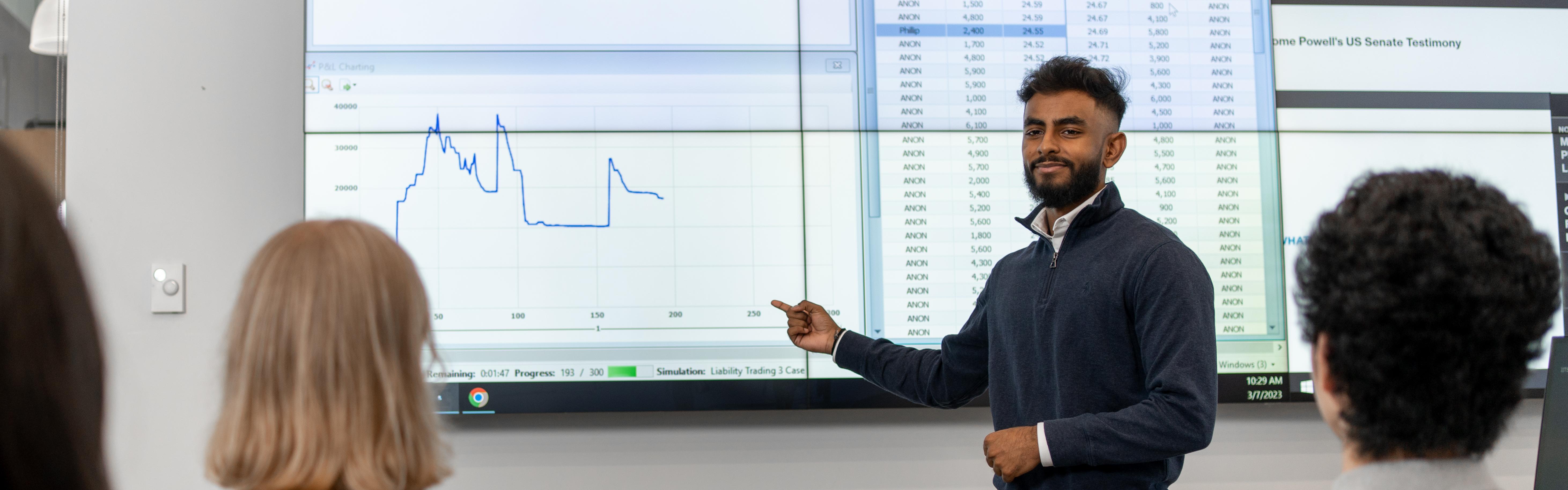 Male student presenting charts and data on a large screen, in front of a classroom of students