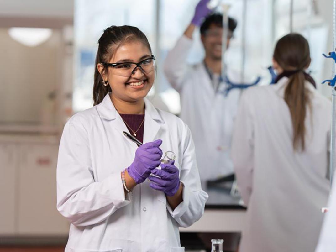 A student in a lab with gloves on holding equipment, smiling