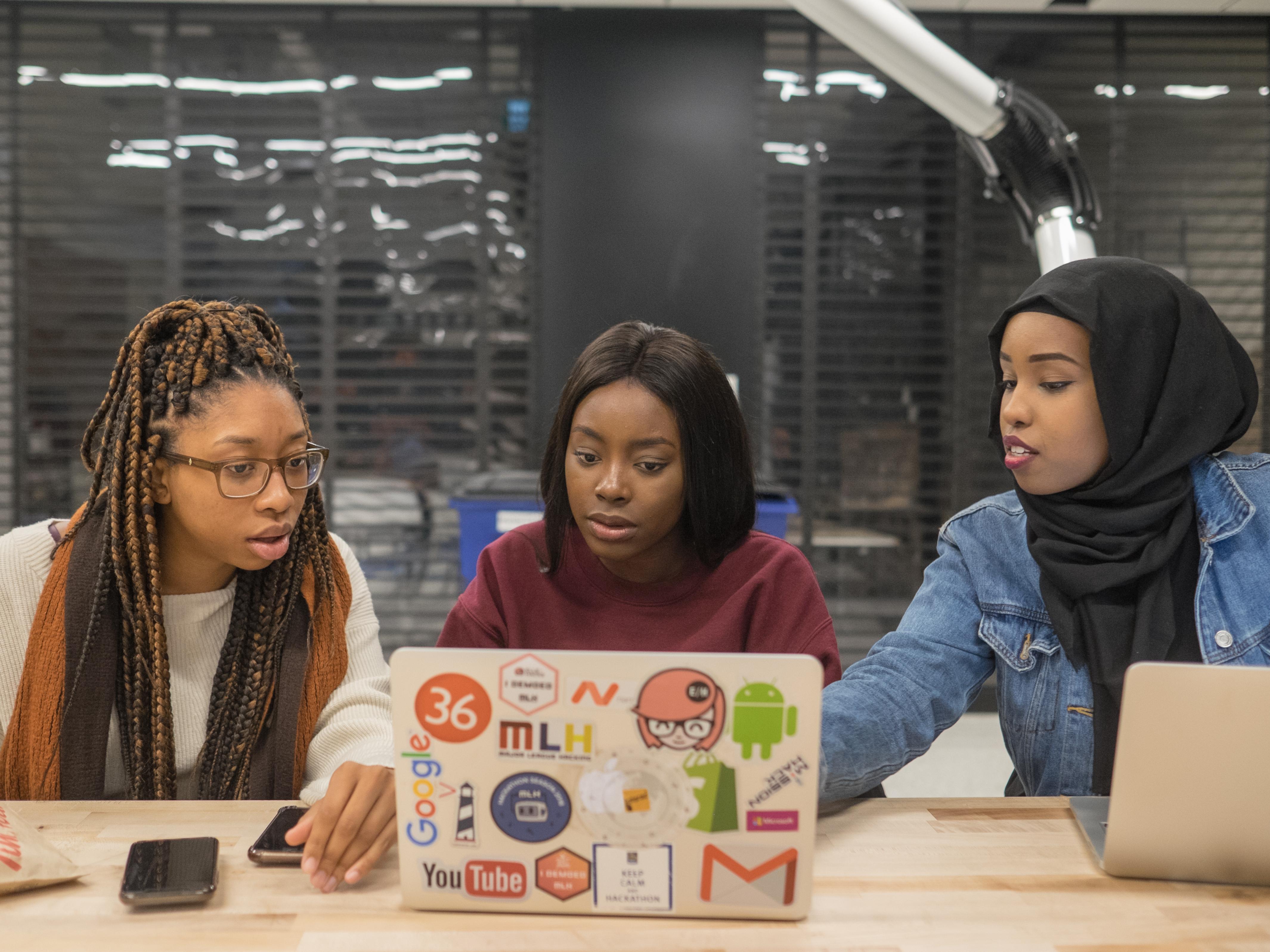 Three dark-skin Black women, one wearing a black Hijab, sit at a desk looking at their laptop computer.