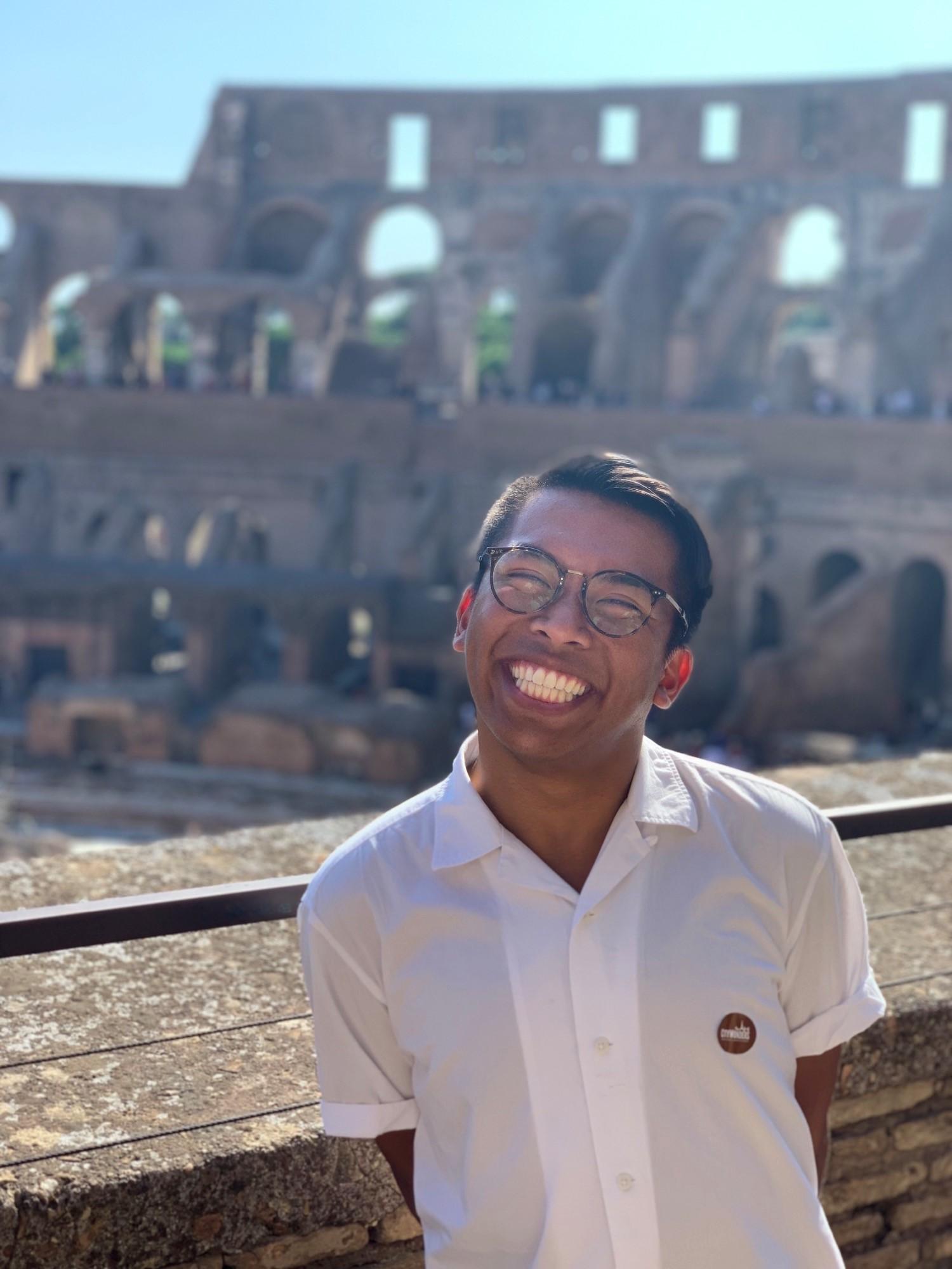 Photo of Alfonso Ralph Mendoza wearing a white short sleeve button down shirt and rounded dark framed glasses in the Colosseum