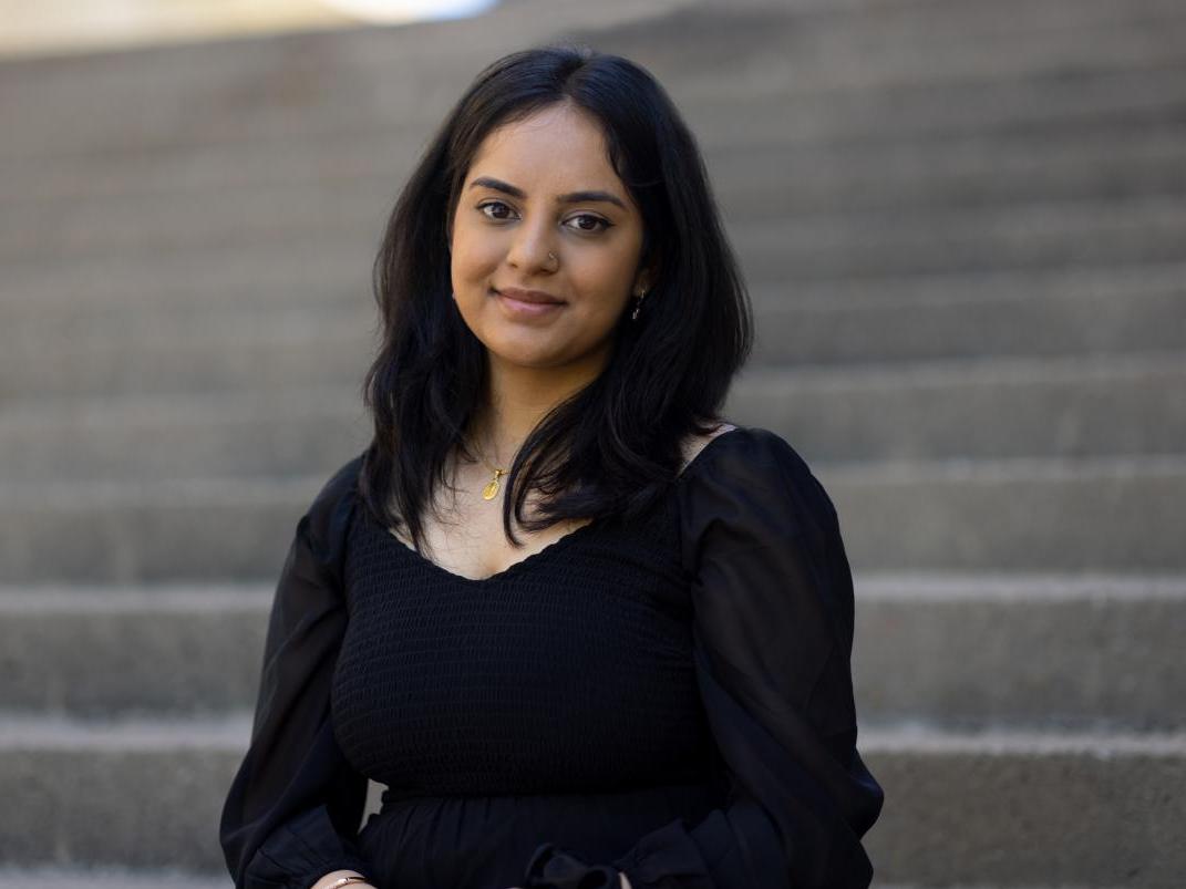 Image of Bhanvi Sachdeva in front of concrete steps with her hair down wearing a gold necklace and black long sleeved top