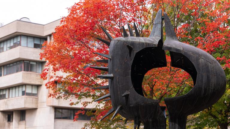 A cropped image of A Tall Couple, a sculpture, in the H-wing patio at the University of Toronto Scarborough, with fall leaves and a building behind