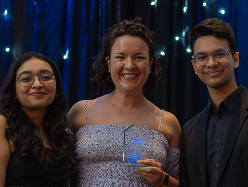 Maria Shibaeva accepts the Professor of the Year Award while smiling widely in front of a starry background with two formally attired students.