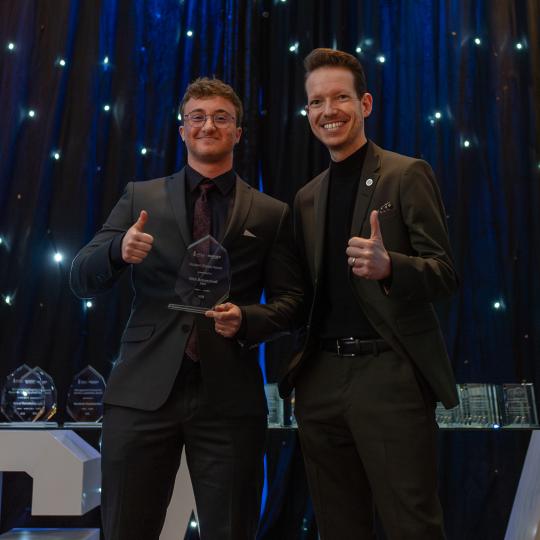 Two men in suits give a thumbs-up while smiling in front of a starry background during the University of Toronto Scarborough Management Gala.