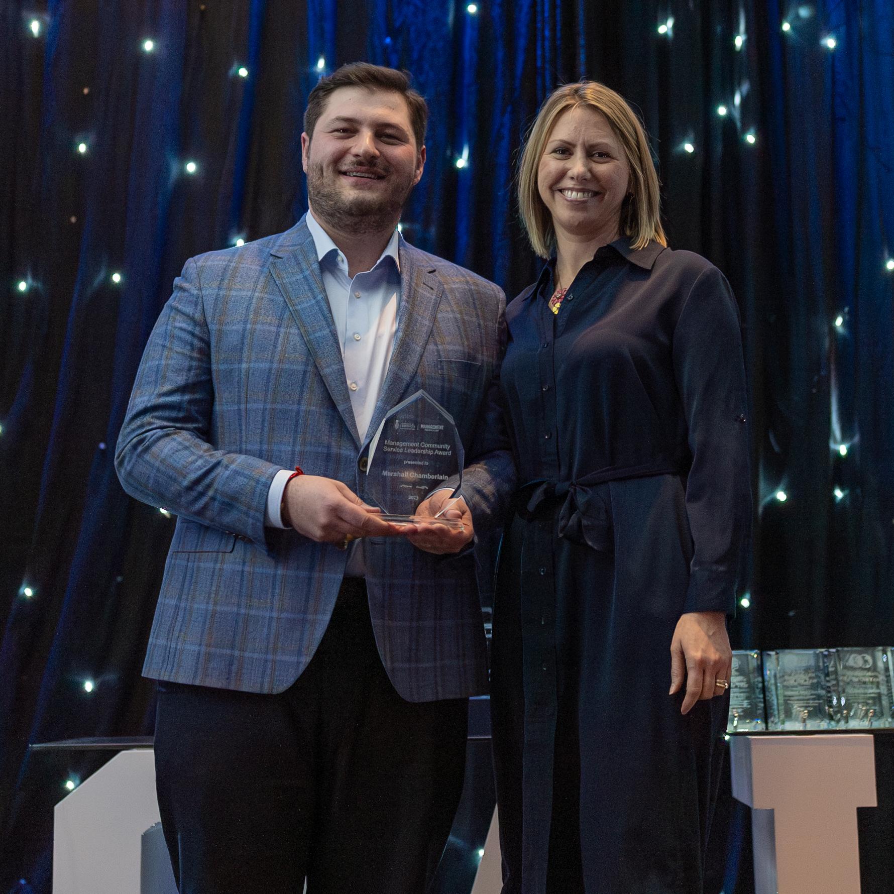 U of T Scarborough Management and International Business alumnus Marshall Chamberlain wearing a plaid jacket holds a glass award in front of a starry background with floor-to-ceiling drapes.