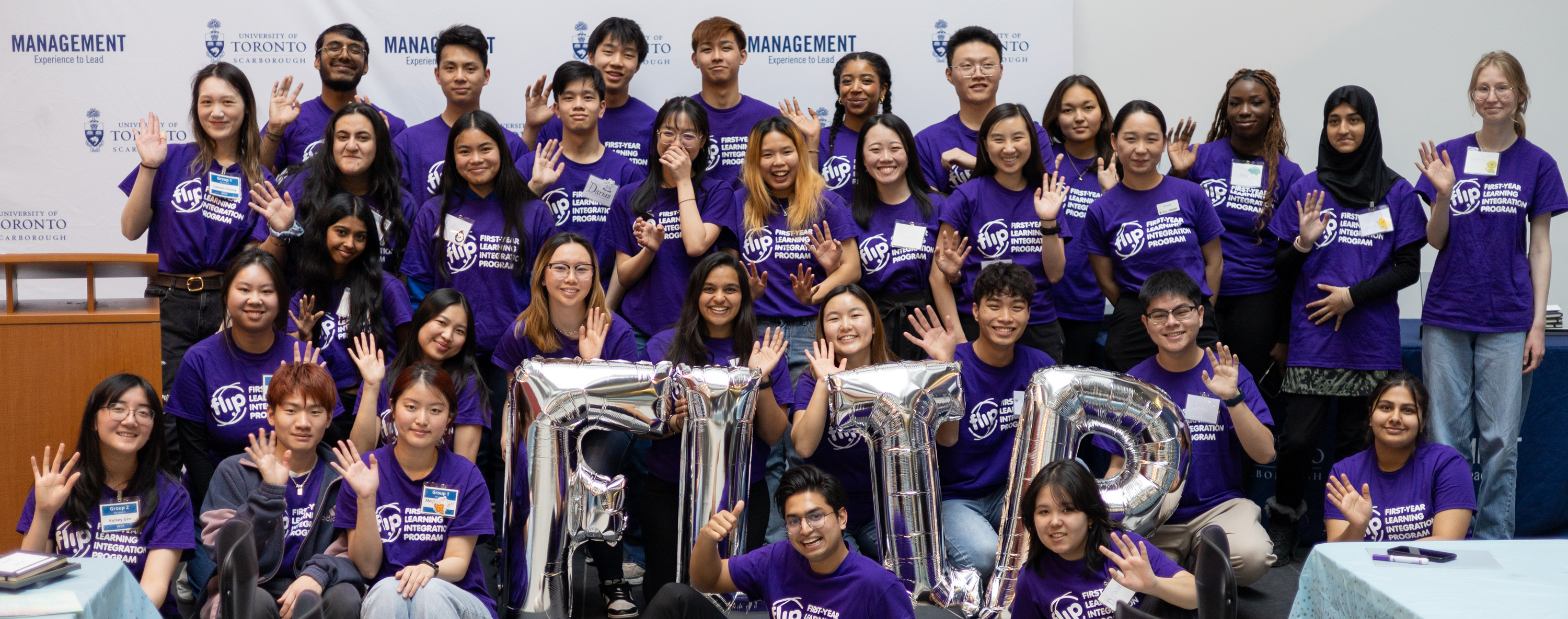 A group of Management students wearing U of T apparel pose for a group photo while smiling widely in the Instructional Centre at the University of Toronto Scarborough with silver balloons that spell "Flip" visible in the foreground.