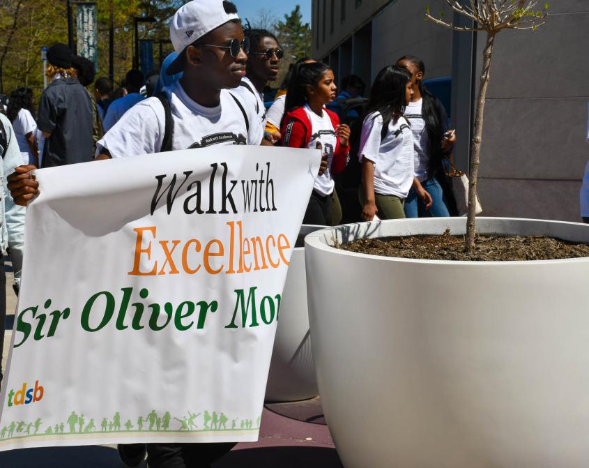 Student from Sir Oliver Mowat Collegiate Institute carries a Walk With Excellence sign as students enter U of T Scarborough. 