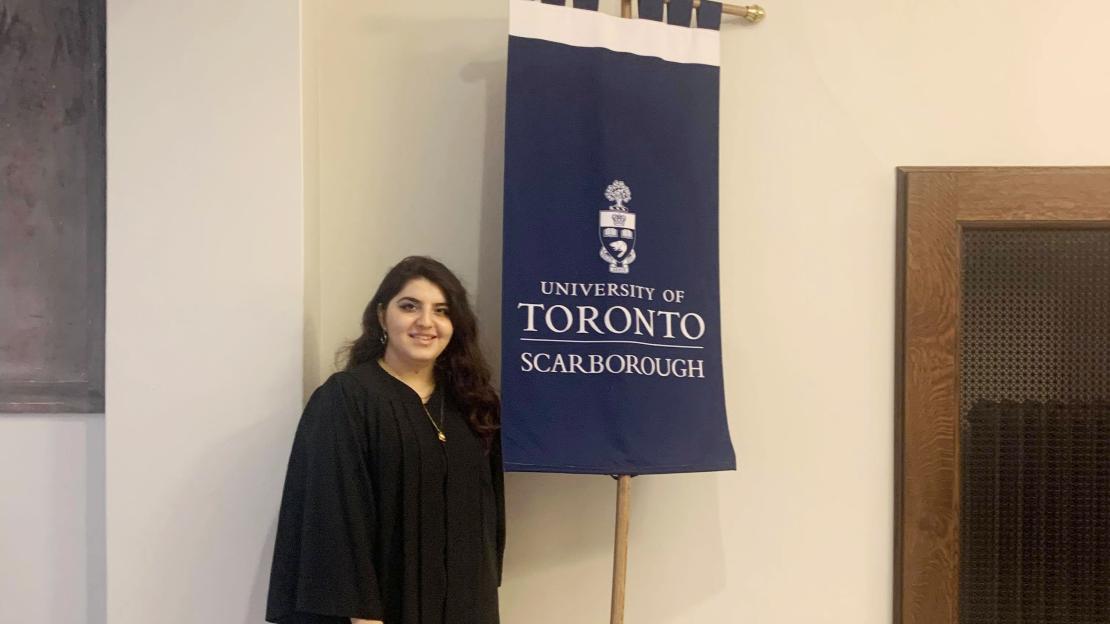 UTSC Student Annie Sahagian stands holding U of T Scarborough gonfalon flag, wearing a black convocation gown.
