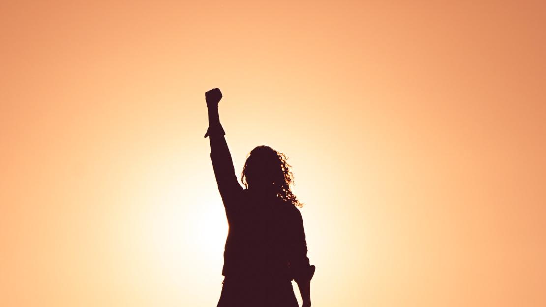 The silhouette of a woman standing with her fist in the air, backlit by a sunset