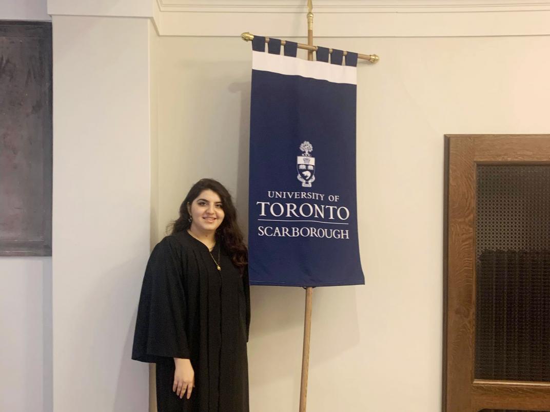 UTSC Student Annie Sahagian stands holding U of T Scarborough gonfalon flag, wearing a black convocation gown.
