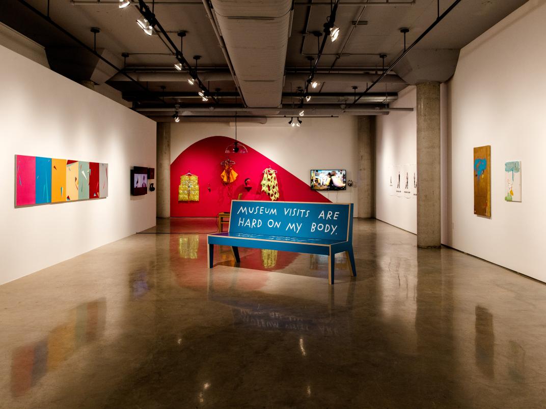 An image of the #CripRitual gallery at the Doris McCarthy Gallery. A blue bench sits in the middle reading "MUSEUM VISITS ARE HARD ON MY BODY"