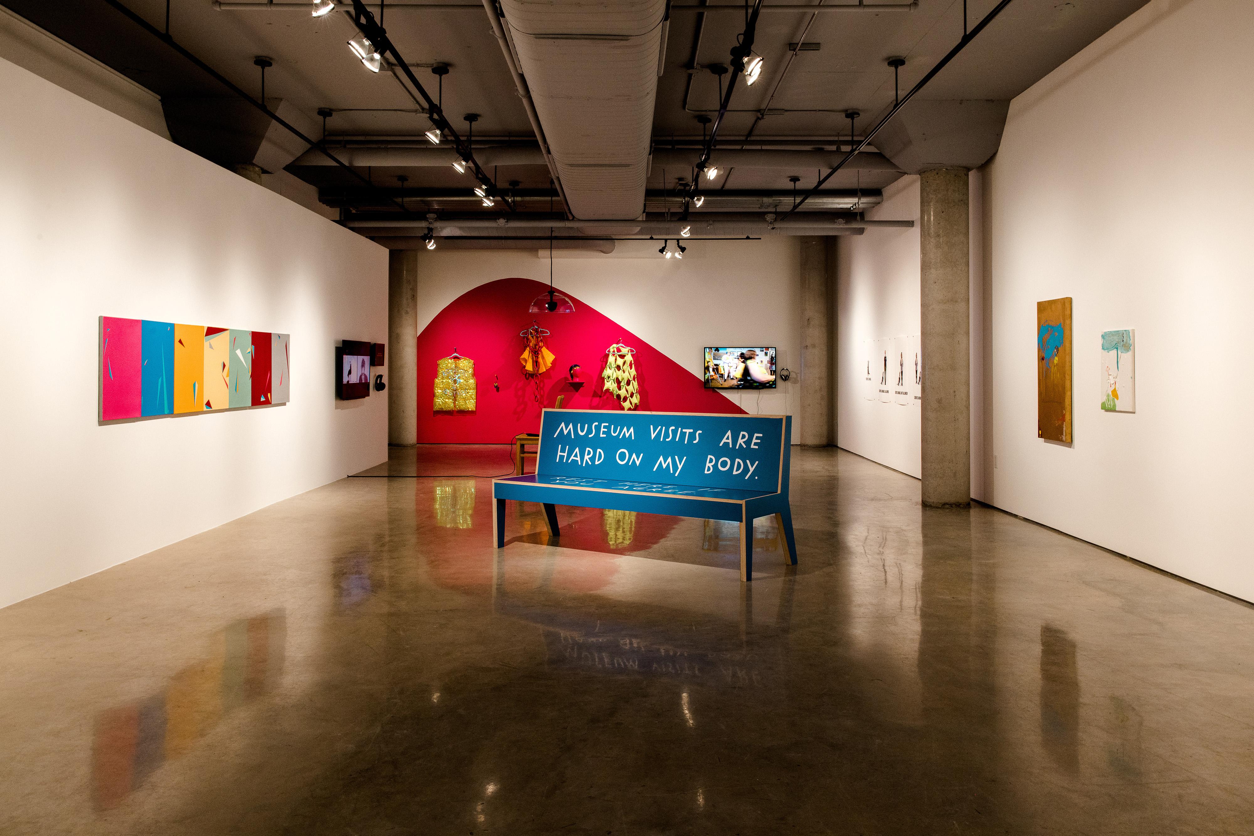 An image of the #CripRitual gallery at the Doris McCarthy Gallery. A blue bench sits in the middle reading "MUSEUM VISITS ARE HARD ON MY BODY"