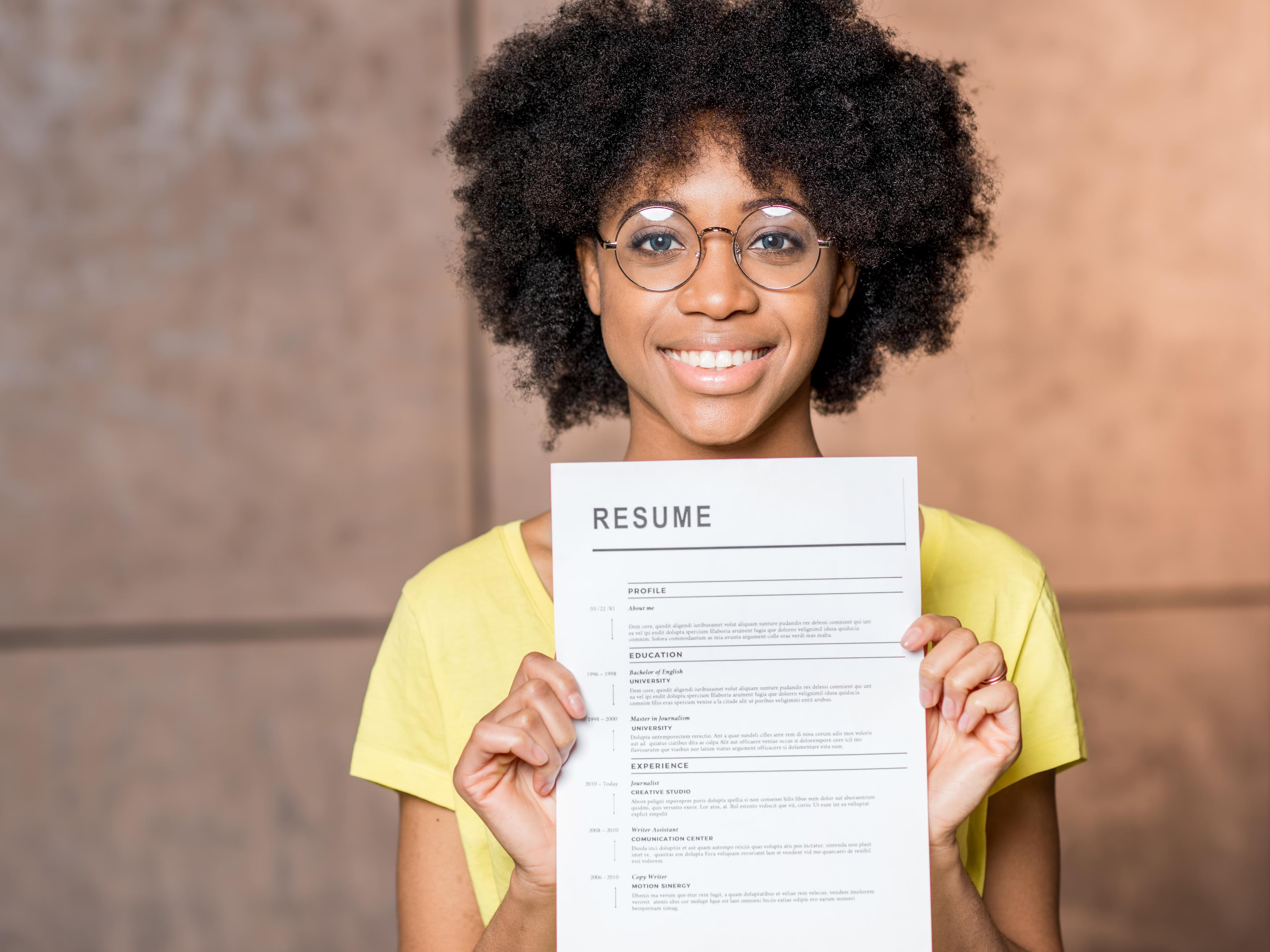 A young black woman with afro hairstyle and round spectacles, smiling and holding up her resume