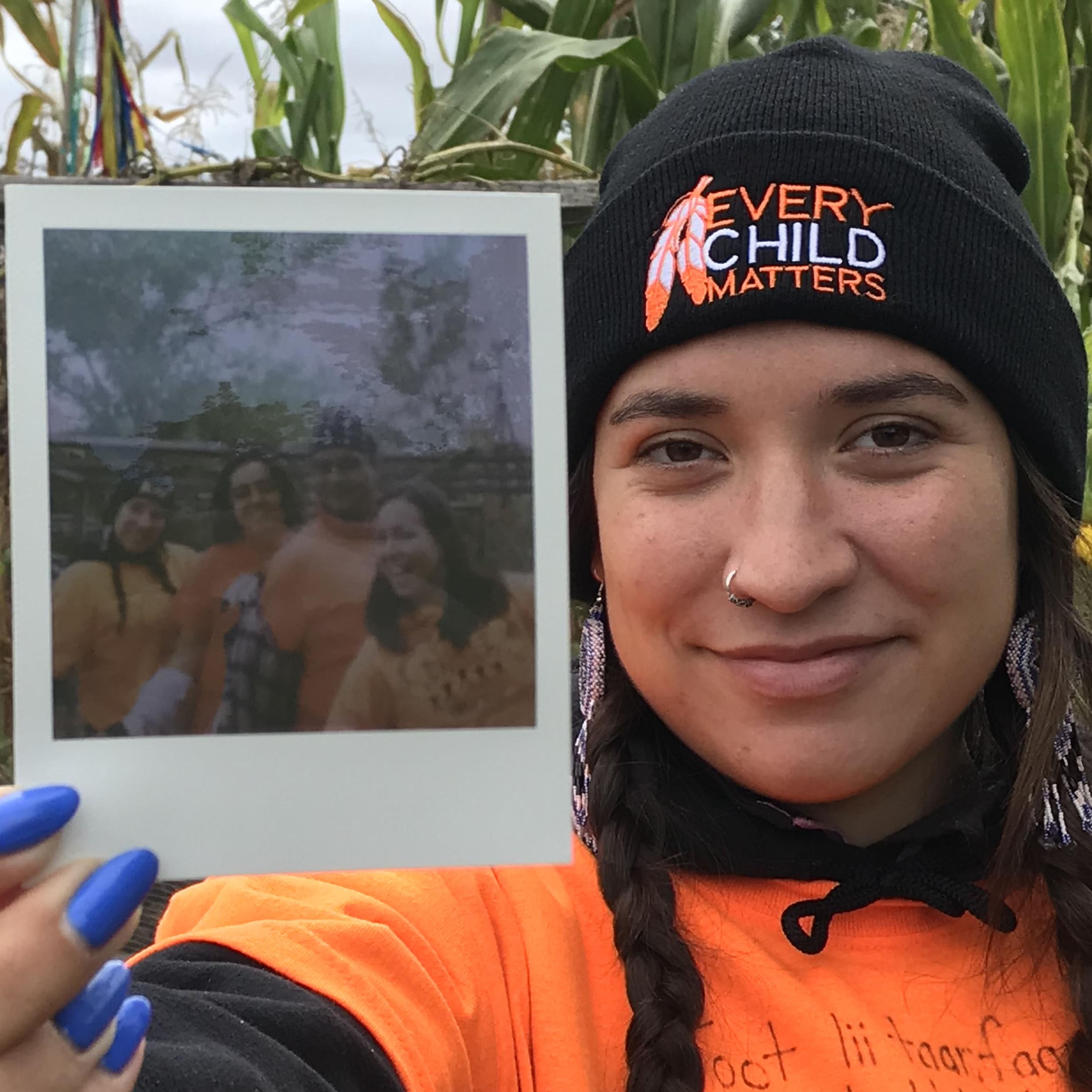 Alexis Bornyk, a young woman with long braided dark hair wearing an "every child matters" hat and orange shirt, holding a polaroid picture from an event at UTSC campus farm