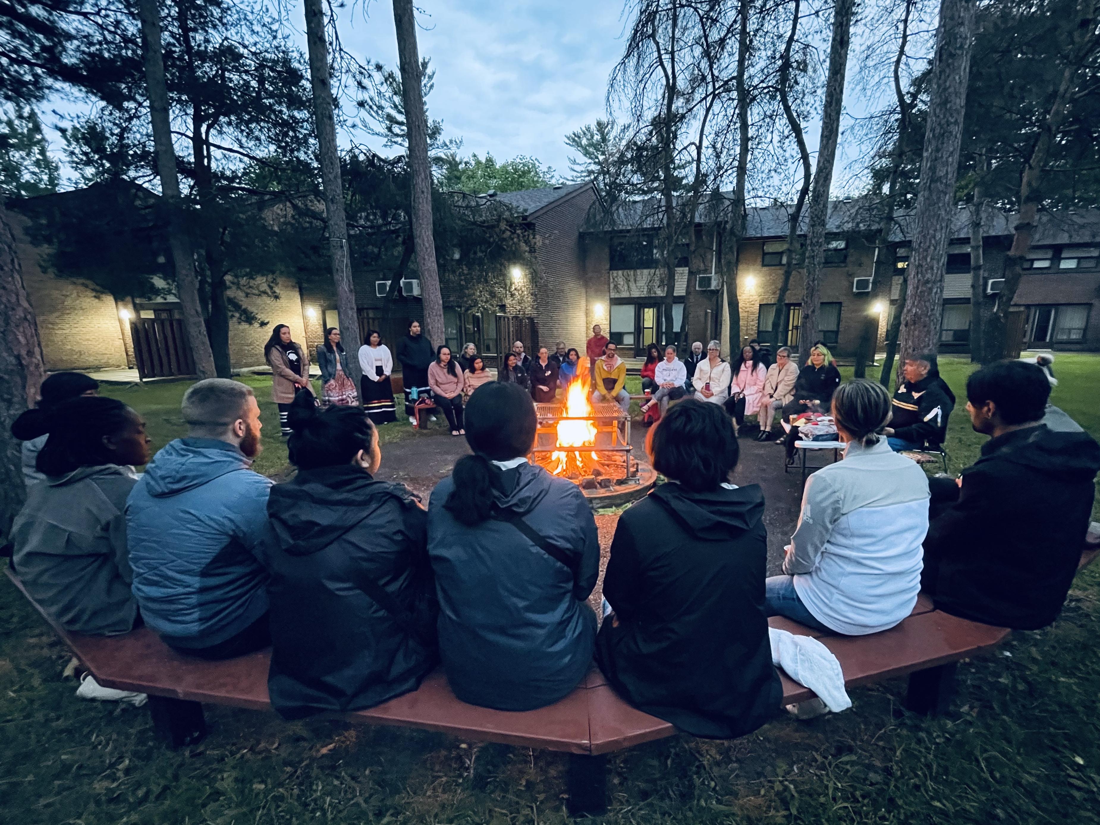 Working Circle Members and guests sitting around a bonfire in front of south residences as sun begins to rise